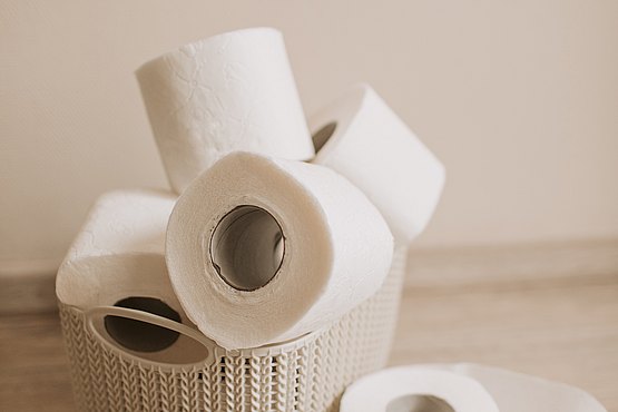Stilisierte Darstellung von Toilettenpapier (Mehrere Rollen von Toilettenpapier in und auf einem kleinem Bast Korb vor hellbraunem Hintergrund.)