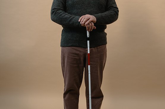 Stilisiertes Bild eines blinden Menschen (Ein älterer Mann mit schwarzer Brille und Blindenstock in der Hand. Gekleidet in brauner Hose und grünem Pullover. Im Hintergrund ist eine braune Wand zu sehen.)