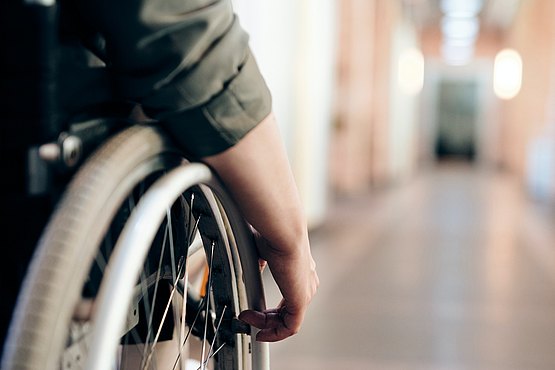 Stilisiertes Bild eines behinderten Menschen (Hochformataufnahme von einem langen hellen Gang. Links im Vordergrund des Bildes ein Mensch im Rollstuhl in grünem Pullover.)