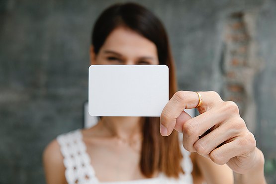 Stilisiertes Bild zur Darstellung eines Angebotes. (Eine junge Frau hält eine weiße, leere Visitenkarte vor Ihr Gesicht - als Angebot eines formalen Kontaktes. Im Hintergrund ist eine graue Wand dargestellt.)