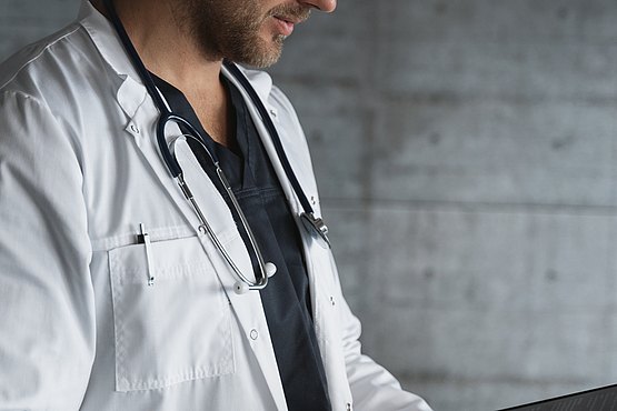 Stilisiertes Bild eines Arztes; 1 junger Arzt in weißem Kittel mit Stethoskop um den Hans gehängt, sowie Klemmbrett in der Hand. Hintergrund als graue Wand.